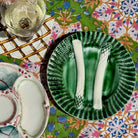 A green ceramic plate with a textured, ribbed rim and two white asparagus spears, styled on a colorful floral tablecloth next to a glass of white wine.