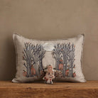 A rectangular beige throw pillow with an embroidered autumn forest scene featuring trees, pumpkins, and a small stitched gingerbread figure, displayed on a wooden surface against a textured beige wall.