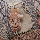 Close-up of a beige fabric textile with embroidered Halloween-themed artwork, including a stitched jack-o'-lantern figure, dark blue tree branches, and autumn leaf patterns.