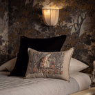 A decorative throw pillow with an embroidered woodland scene featuring a moonlit owl, fox, and pumpkins, styled on a bed with a black velvet pillow and a warm wall lamp in the background.