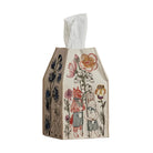 A tissue box cover made of cream-colored paper with a whimsical hand-drawn illustration of a rabbit, fox, and flowers in muted red, blue, and orange tones, sitting on a wooden surface against a textured beige wall.
