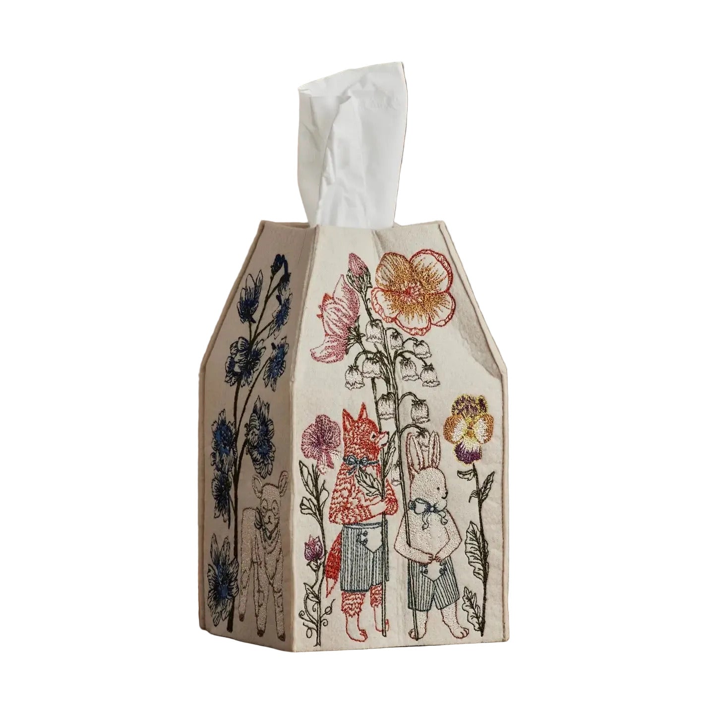 A tissue box cover made of cream-colored paper with a whimsical hand-drawn illustration of a rabbit, fox, and flowers in muted red, blue, and orange tones, sitting on a wooden surface against a textured beige wall.