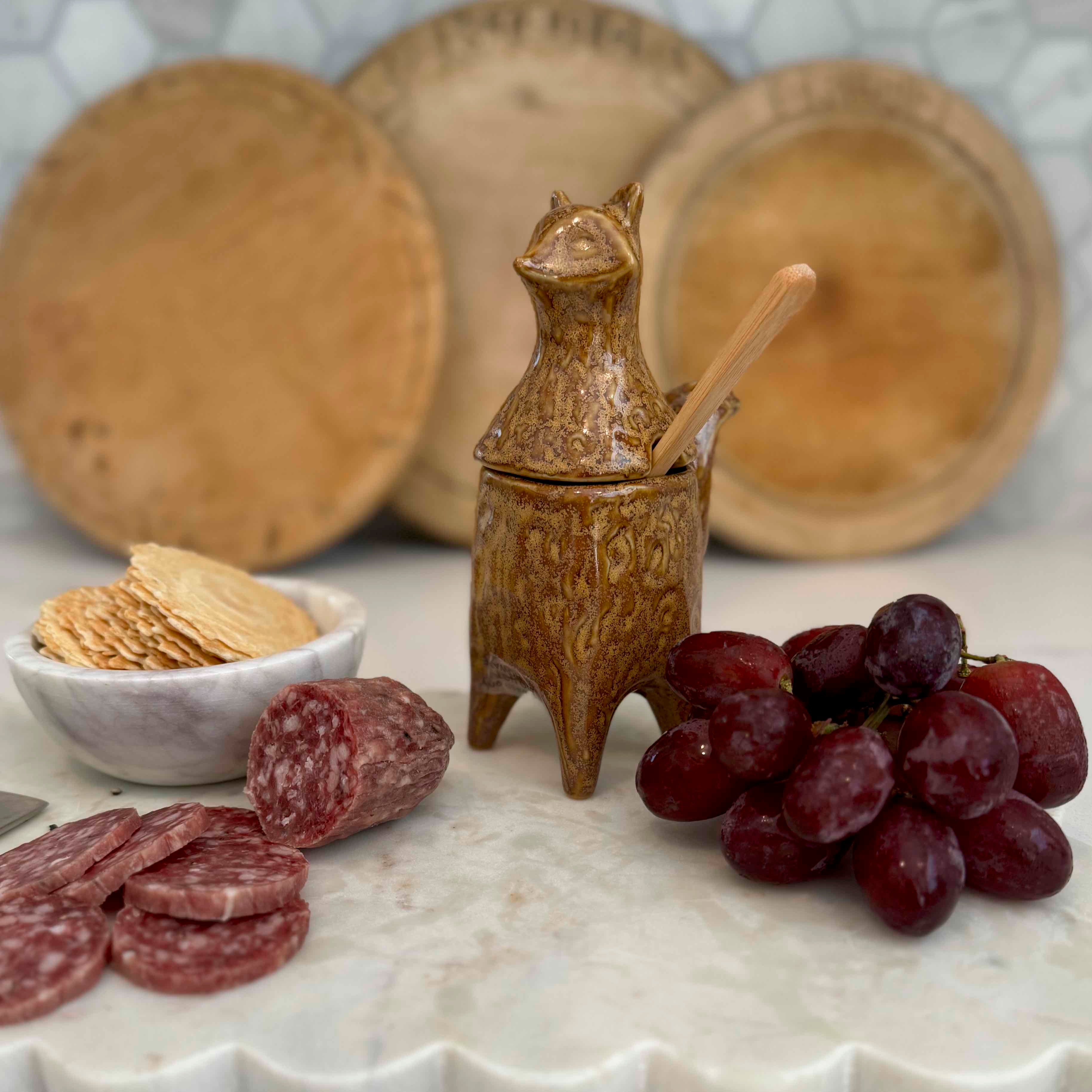 Decorative ceramic container with a spoon, crackers, salami, and grapes on a marble surface.