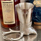 Metallic boot-shaped jigger with bottles of whiskey in the background