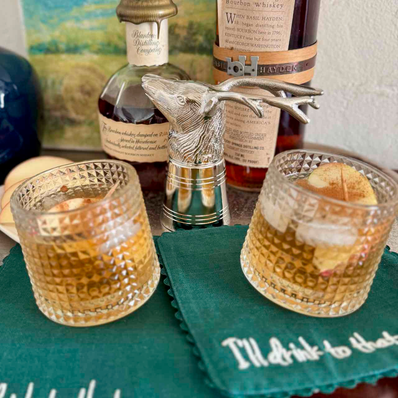 Two glasses of whiskey with a decorative deer head decanter and bottles in the background.