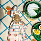 A folded patterned napkin with a cherry and grid print, secured with a clear faceted star-shaped napkin ring, placed on a table setting with green ceramic dishes and food props.