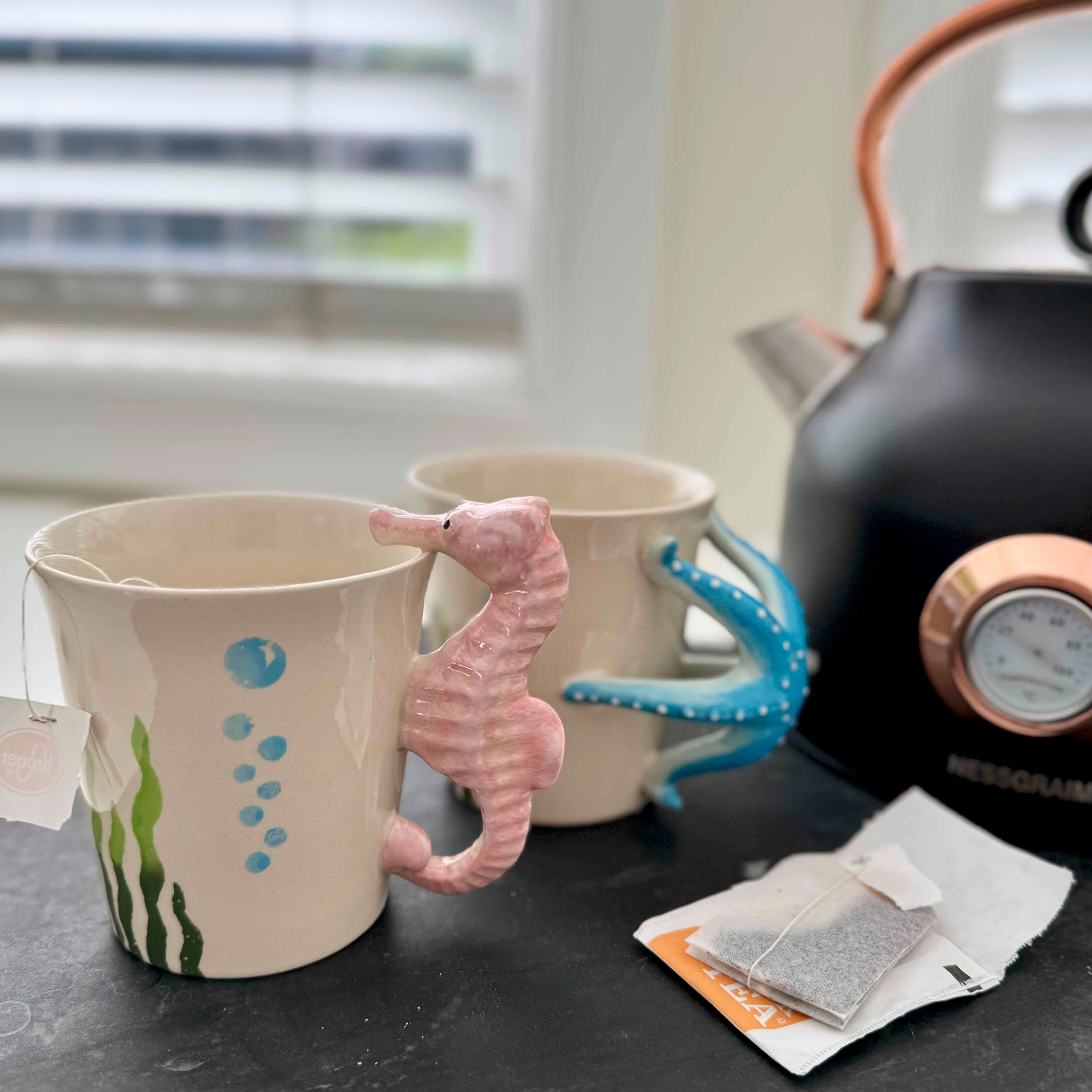 Ceramic mug with seahorse handle on a table with tea bags and a teapot.