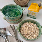 Two green ceramic bowls with soup and oats, a yellow milk carton, and spoons on a white surface.