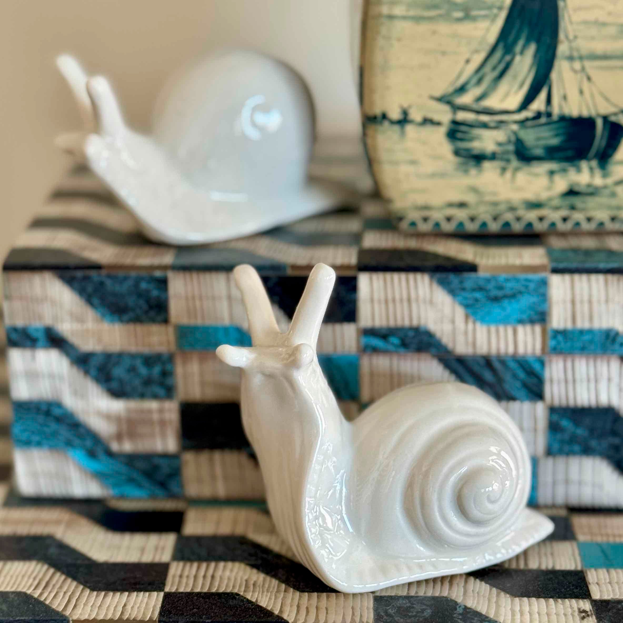 Two ceramic snail figurines on a checkered surface with a sailboat painting in the background.