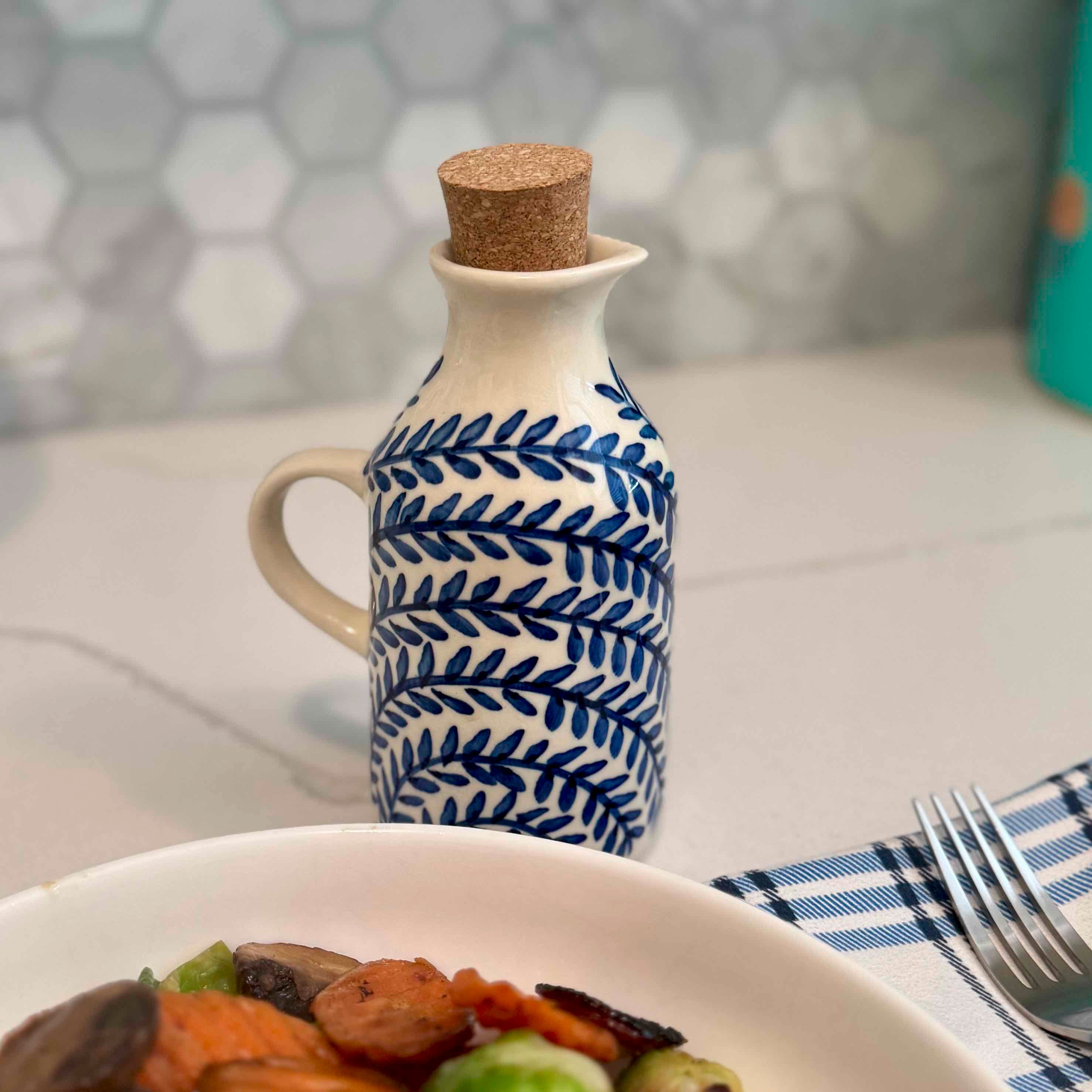 Ceramic oil bottle with blue leaf pattern on a table with a plate of food and cutlery.
