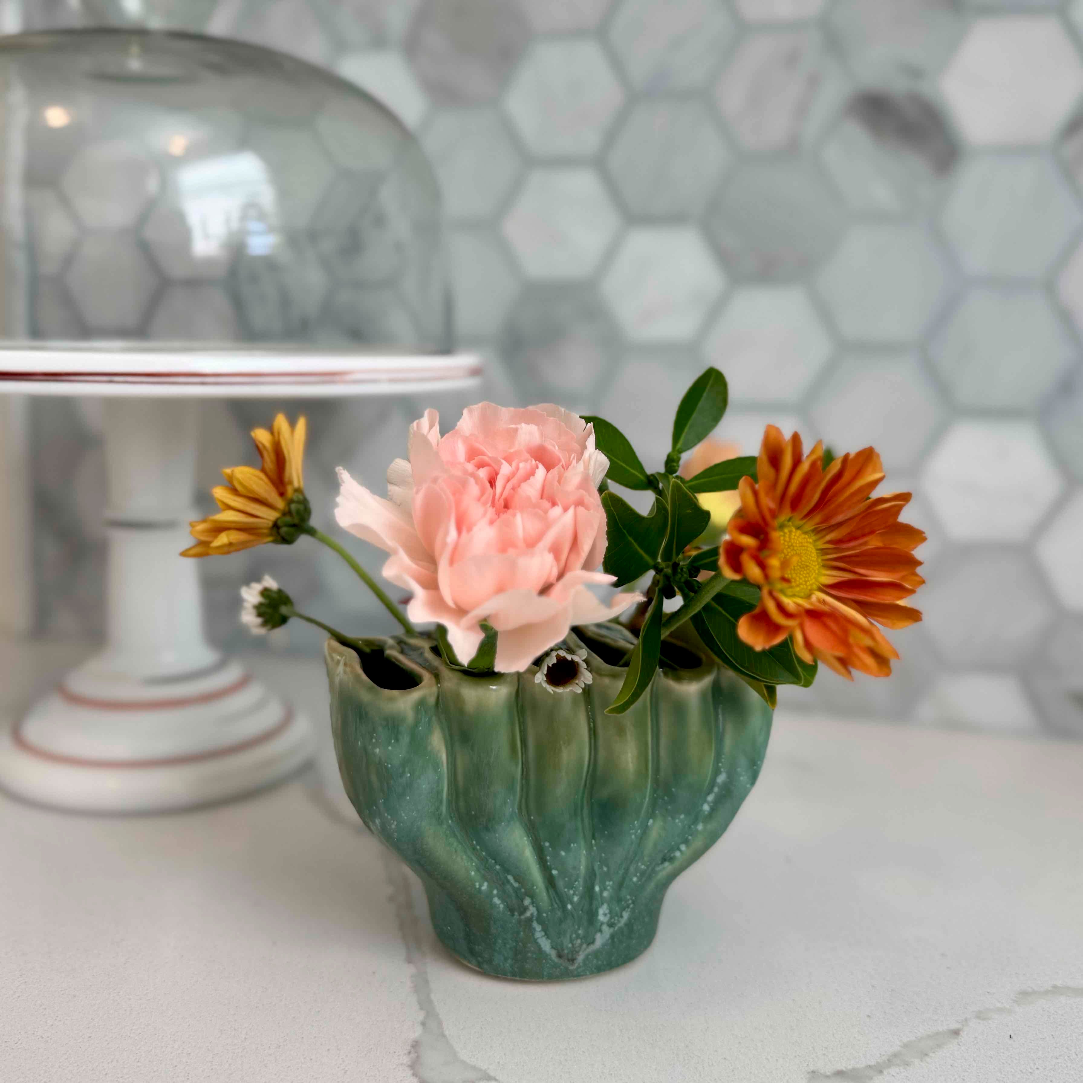 Green vase with pink and orange flowers on a marble surface with a hexagonal patterned wall in the background.