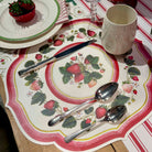Table setting with strawberry-themed placemat, plates, and cutlery on a striped tablecloth.