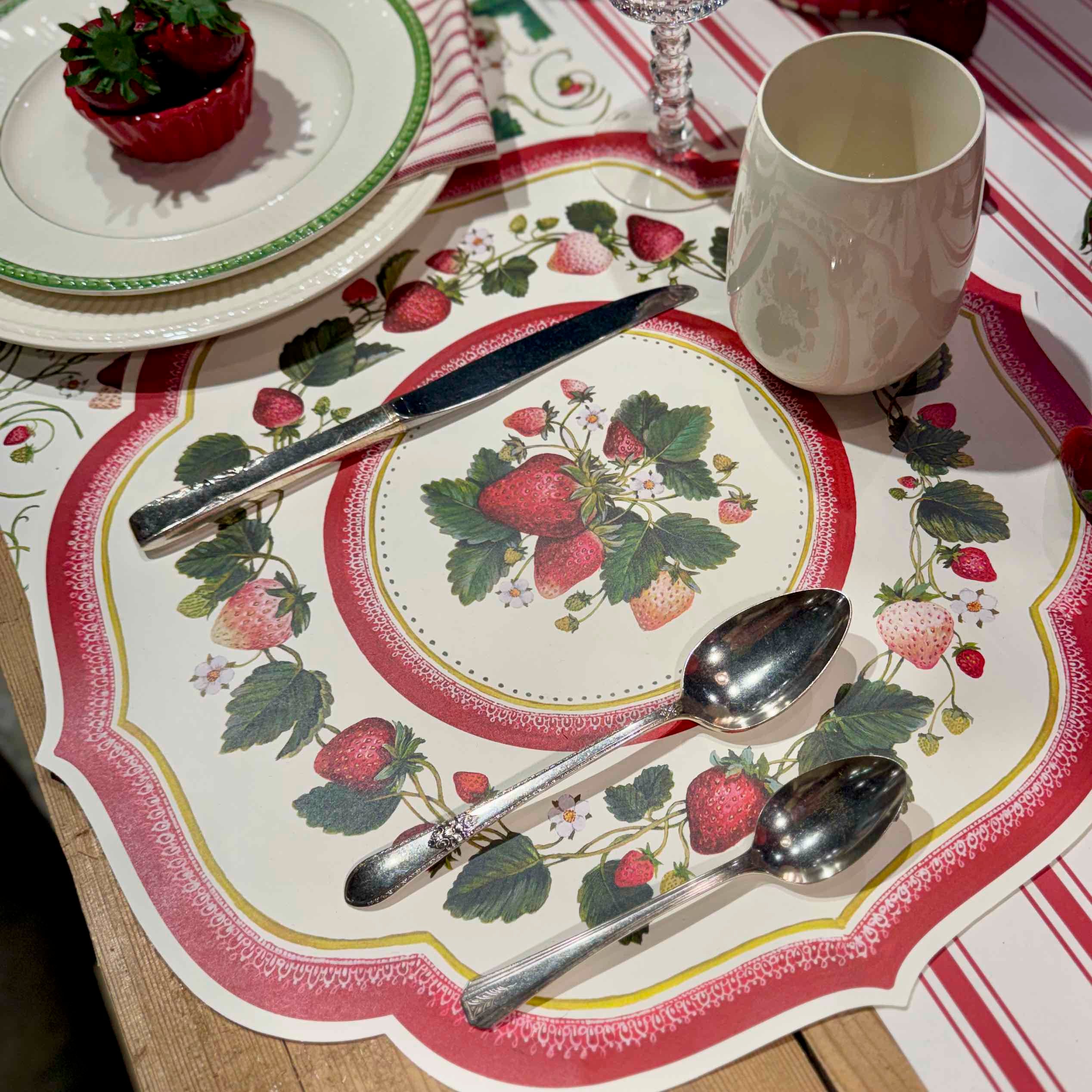 Table setting with strawberry-themed placemat, plates, and cutlery on a striped tablecloth.