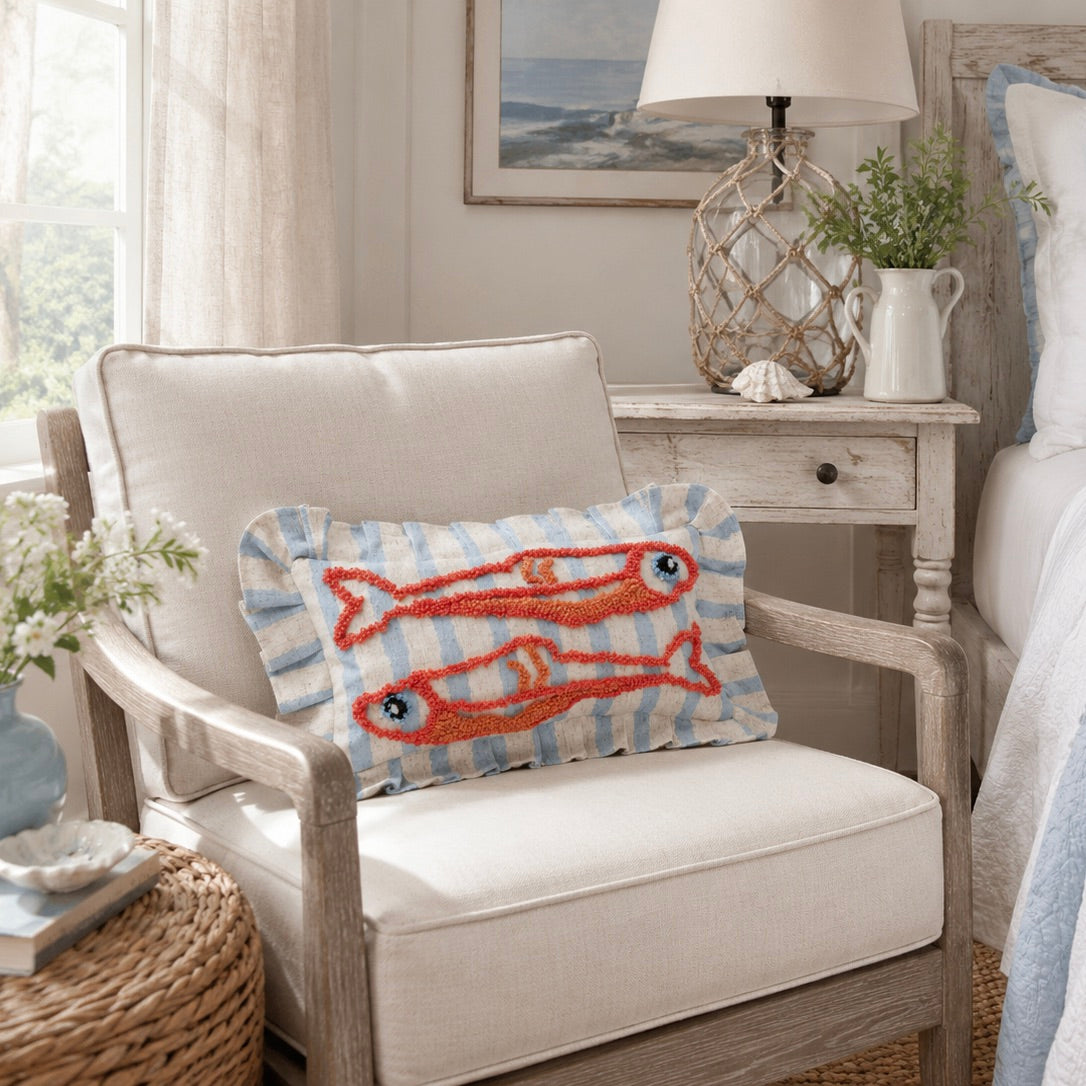 A decorative throw pillow featuring two orange-red embroidered fish on a light blue and white striped background, styled on a cream-colored armchair in a bright, coastal-themed bedroom setting.