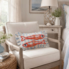 A decorative throw pillow featuring two orange-red embroidered fish on a light blue and white striped background, styled on a cream-colored armchair in a bright, coastal-themed bedroom setting.