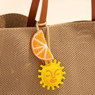 Close-up of a woven straw tote bag with brown handles, featuring a hanging beaded charm shaped like a yellow sun and an orange slice.