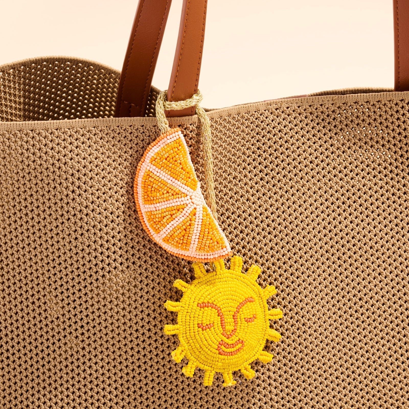Close-up of a woven straw tote bag with brown handles, featuring a hanging beaded charm shaped like a yellow sun and an orange slice.