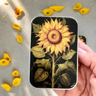Phone case with sunflower design held by a hand on a gray surface with scattered petals.