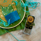 A close-up of sewing and embroidery tools on a wooden surface, including a green fabric hoop with floral embroidery, a blue felt sample, turquoise thread, and a small metal tin with a sunflower illustration and pins.