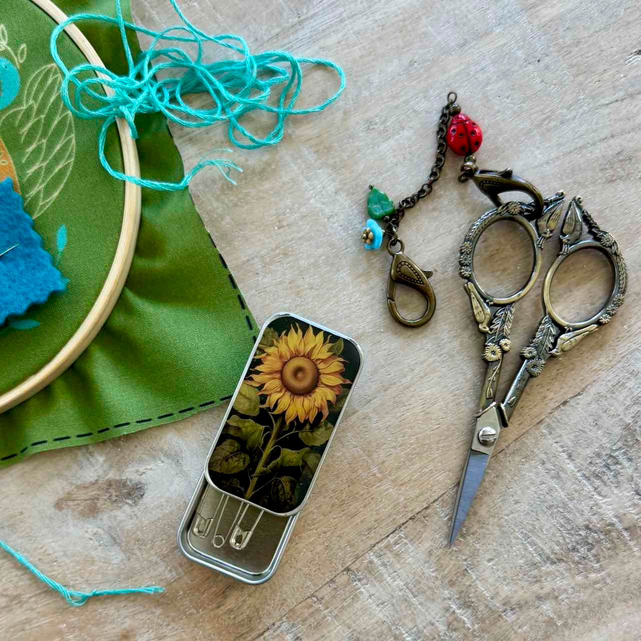A styled flat-lay of sewing and crafting tools on a light wood surface, including ornate silver scissors, a small tin with a sunflower illustration and safety pins, turquoise thread, a green fabric hoop, and a decorative charm.