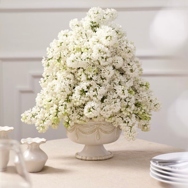 A large arrangement of dense white baby's breath flowers in an ornate cream-colored pedestal vase, set on a light beige tabletop with decorative ceramic vases and stacked plates in the background.