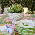 A stack of white ceramic plates with green rims and a leaf motif, arranged on a colorful floral tablecloth next to a white pedestal planter holding a green succulent, with an outdoor garden background.