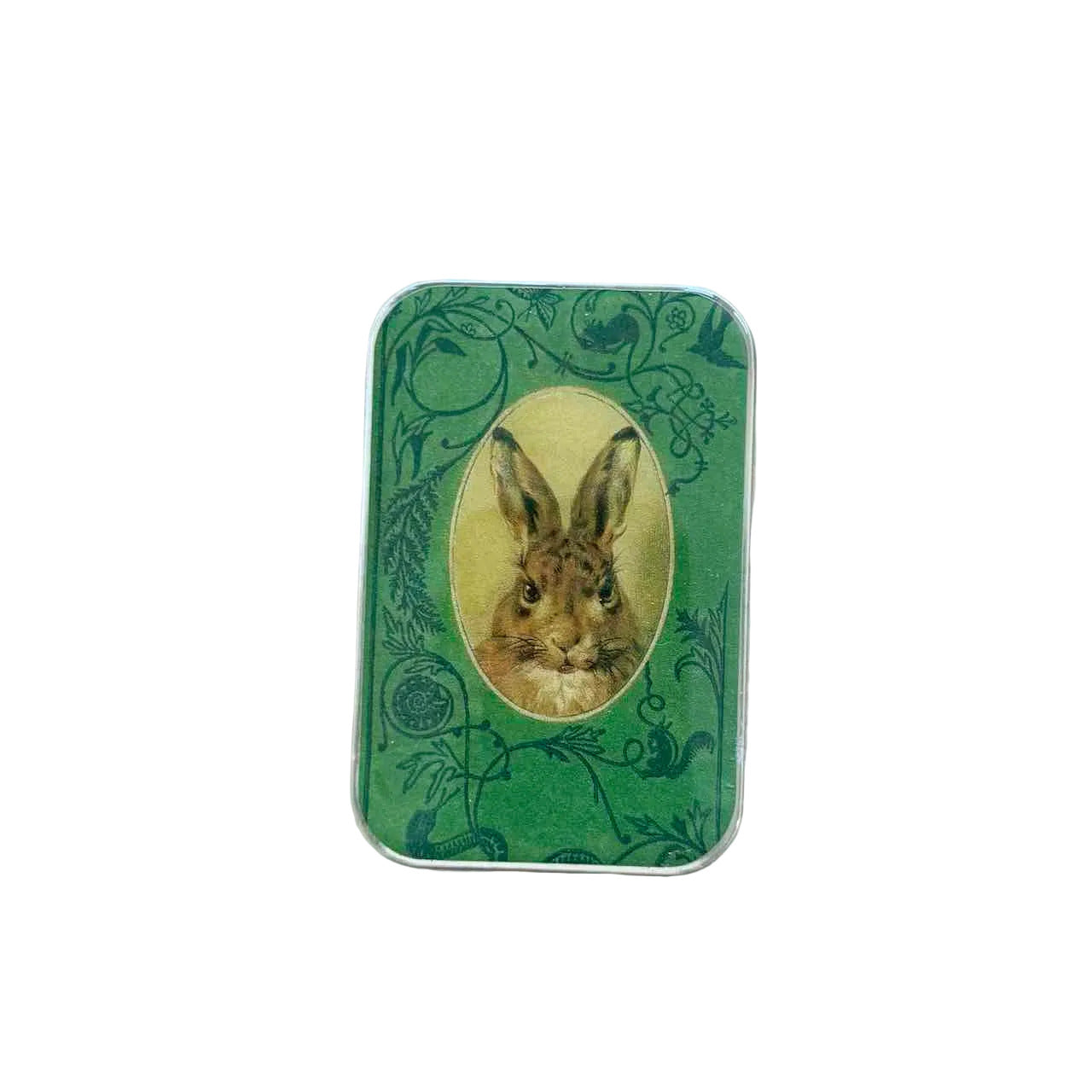 A rectangular green tin with rounded corners, featuring a central oval illustration of a brown rabbit surrounded by dark green floral and vine patterns, placed on a light wood surface.