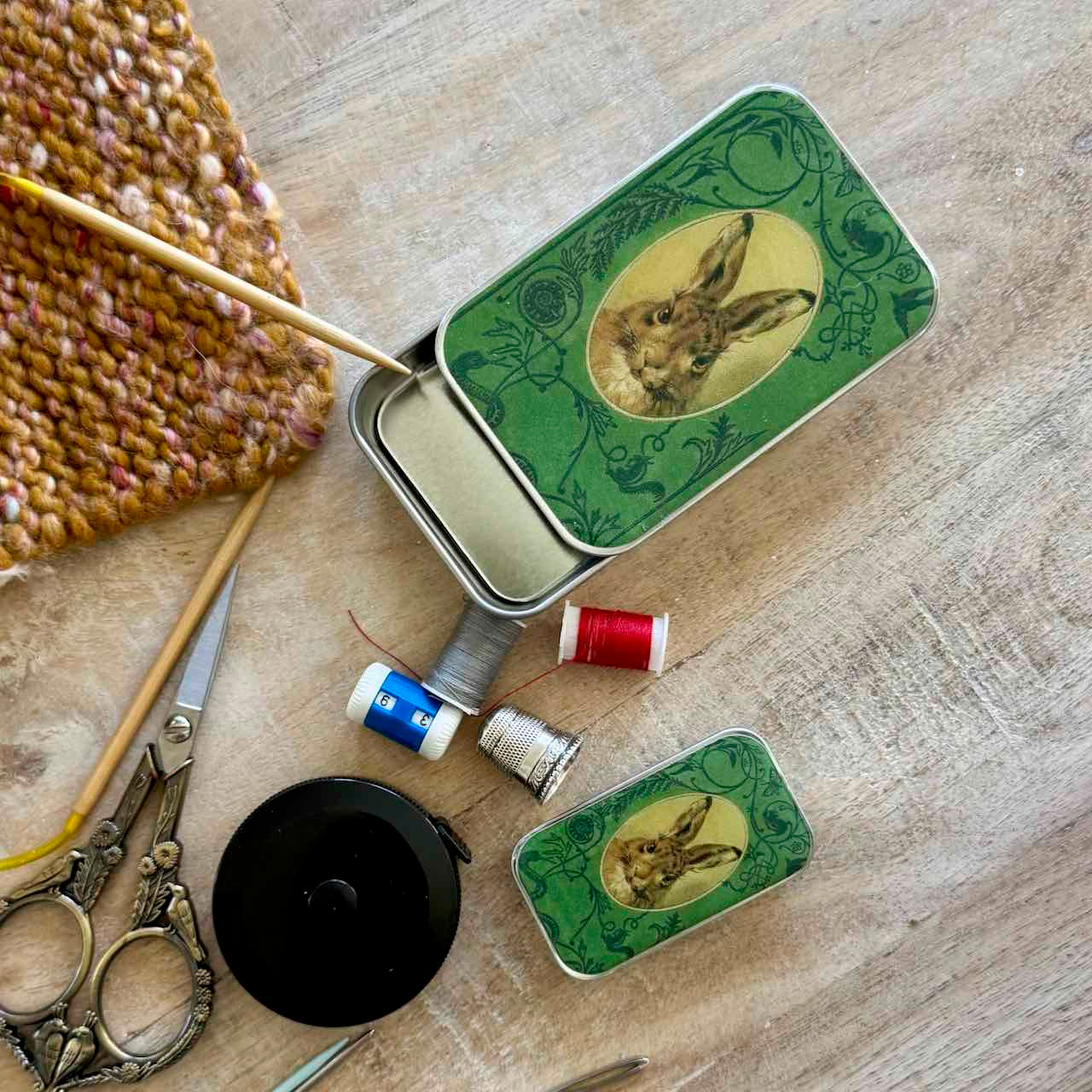 A styled flat-lay of vintage-style metal sewing pincases with green rabbit illustrations, shown alongside sewing tools including scissors, thread spools, a black tape measure, and a knitted fabric on a light wood surface.