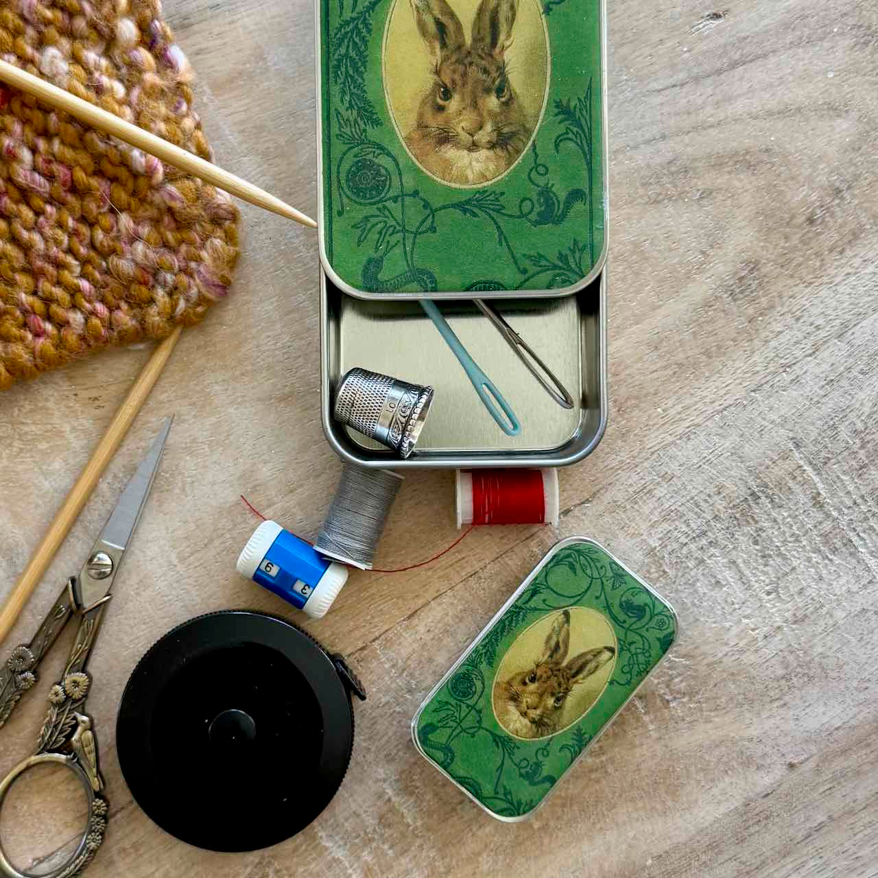 A styled flat-lay of a green metal sewing kit tin with a rabbit illustration, shown open with thread spools, a thimble, pins, and a needle on a wooden surface, alongside knitting tools and scissors.