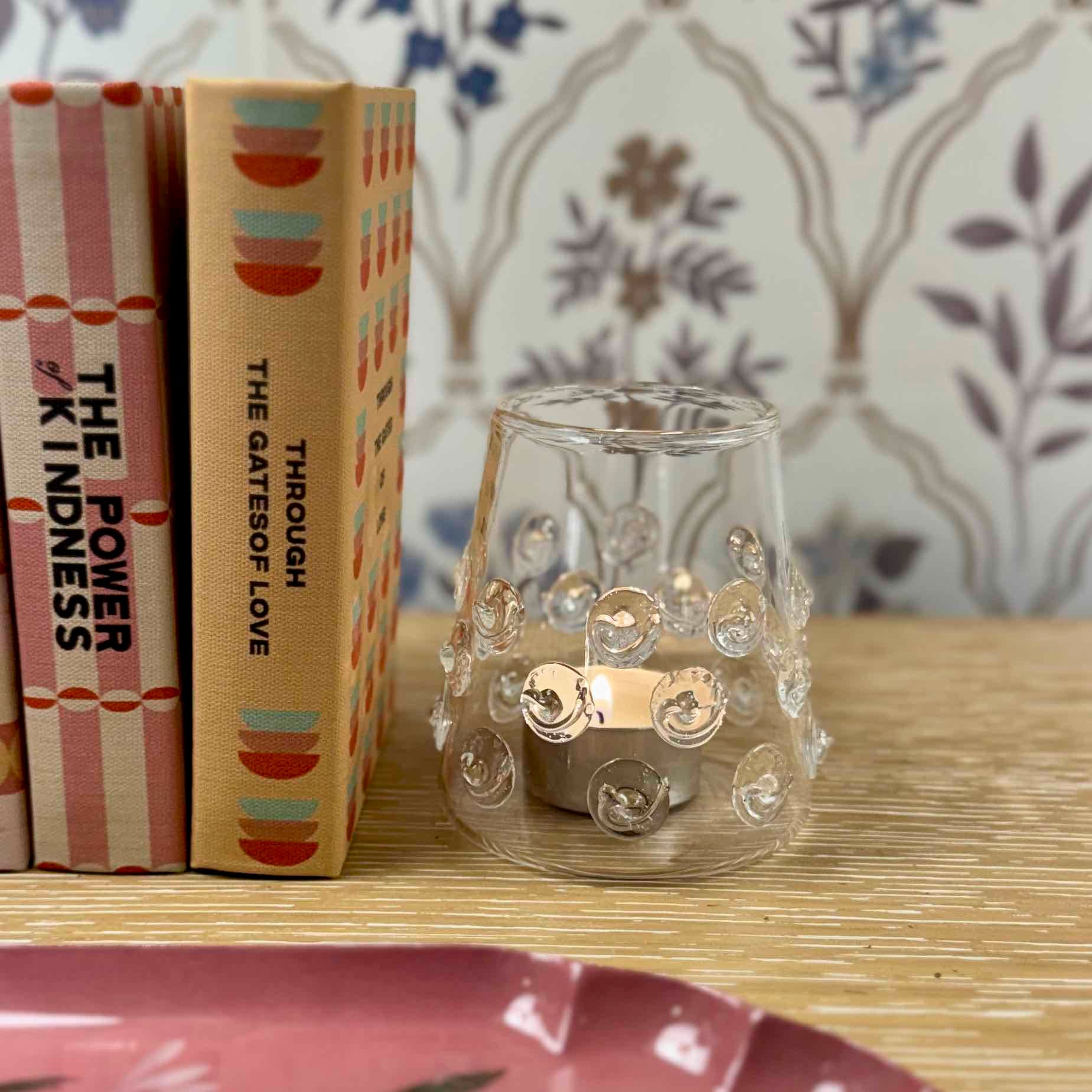 Clear glass candle holder with decorative elements on a wooden surface with books in the background.