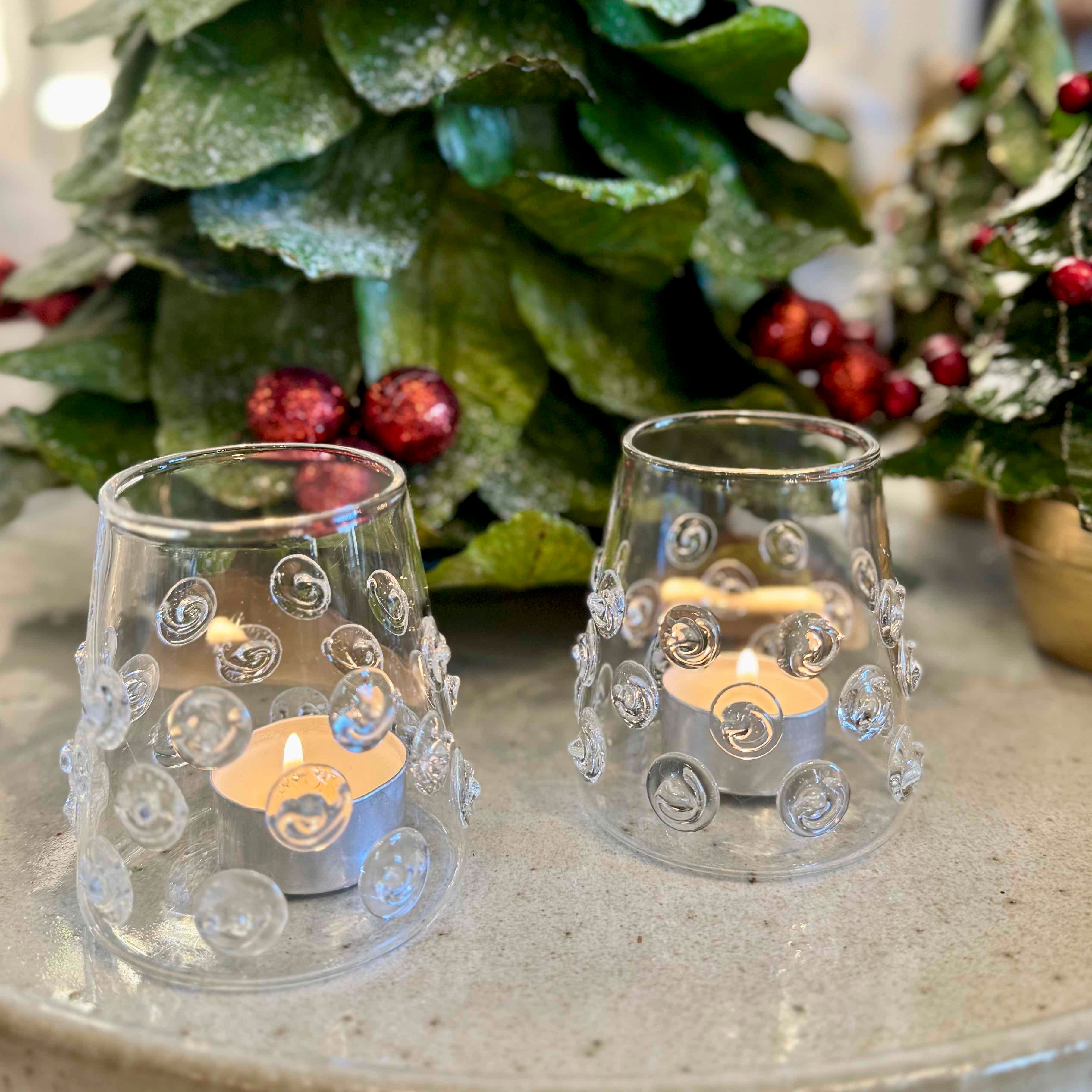 Two clear glass candle holders with decorative patterns on a surface with a Christmas tree in the background.