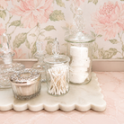 Decorative glass jars with cotton swabs and a silver container on a marble surface with floral wallpaper.