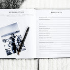 Open book with family tree and basic facts questionnaire on a wooden surface with a pen and photo.