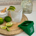 Cocktail preparation with limes, a glass, and a green ice cube tray on a wooden board.