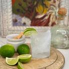 Frosted glass with lime garnish on a wooden coaster with limes and a tequila bottle in the background.