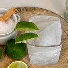 Clear glass with lime wedges on a wooden surface next to a jar of salt.