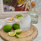 Lime wedges and a glass with ice on a wooden board, with a bottle in the background.