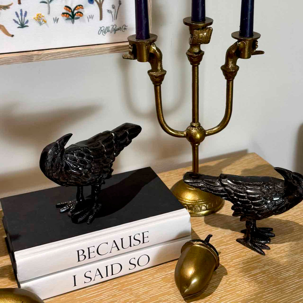 Two black raven figurines displayed on a wooden table, one standing on a black book and the other beside a gold candelabra and acorn decor.