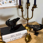 Two black raven figurines displayed on a wooden table, one standing on a black book and the other beside a gold candelabra and acorn decor.