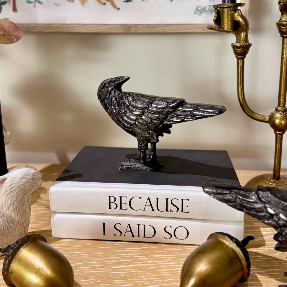 A dark metallic raven figurine displayed on top of two stacked books that read 'BECAUSE I SAID SO', with decorative brass objects and part of another bird visible around it.