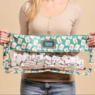 A smiling young woman holds up a clear zippered pouch filled with small white cards or blocks, featuring a teal and cream patterned fabric top with the brand name 'Madison Tiles' visible on the front label.