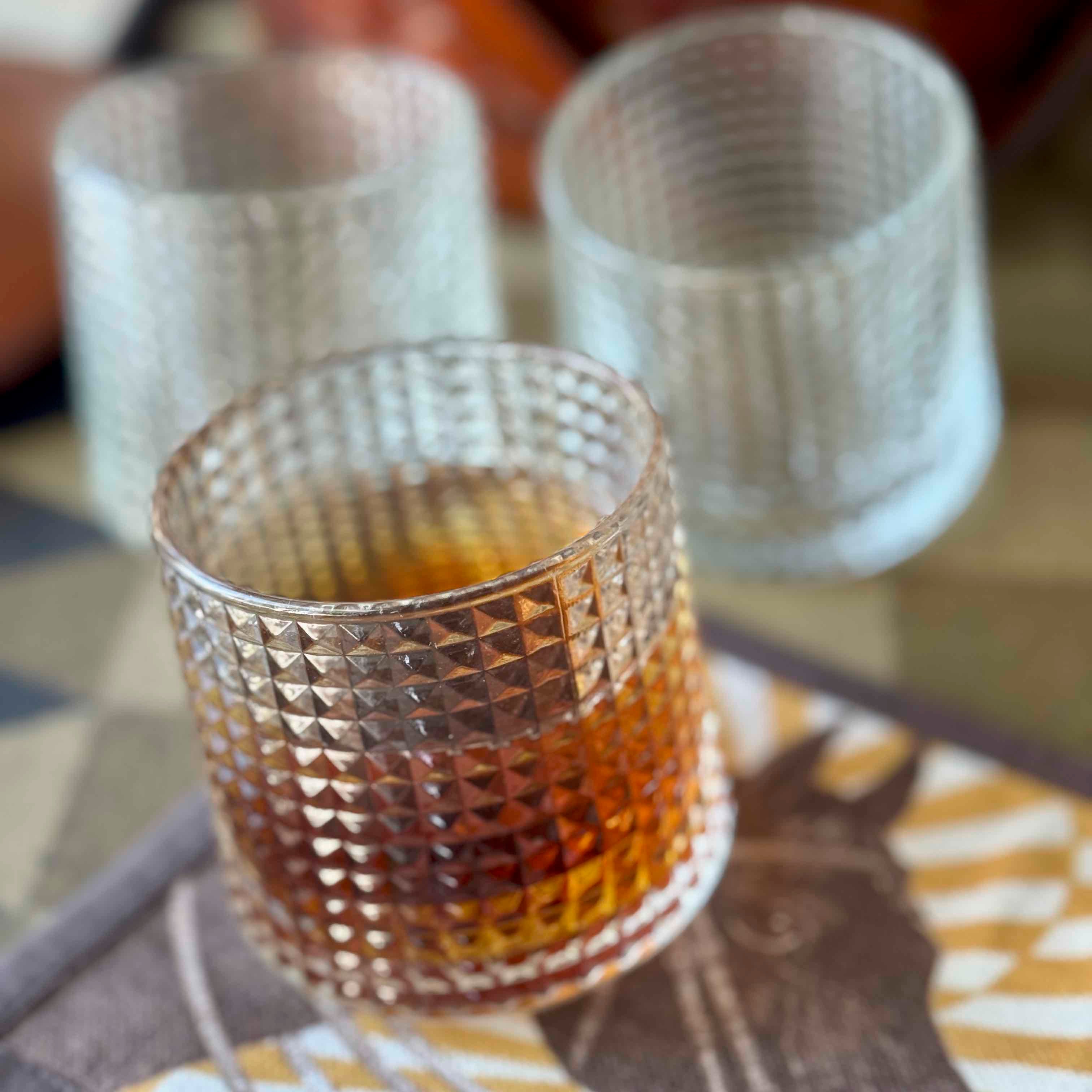 Decorative glass with textured pattern containing a amber liquid, placed on a surface with blurred background.