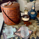 Brown woven leather basket with two bottles of alcohol, glasses, and tools on a checkered tablecloth.