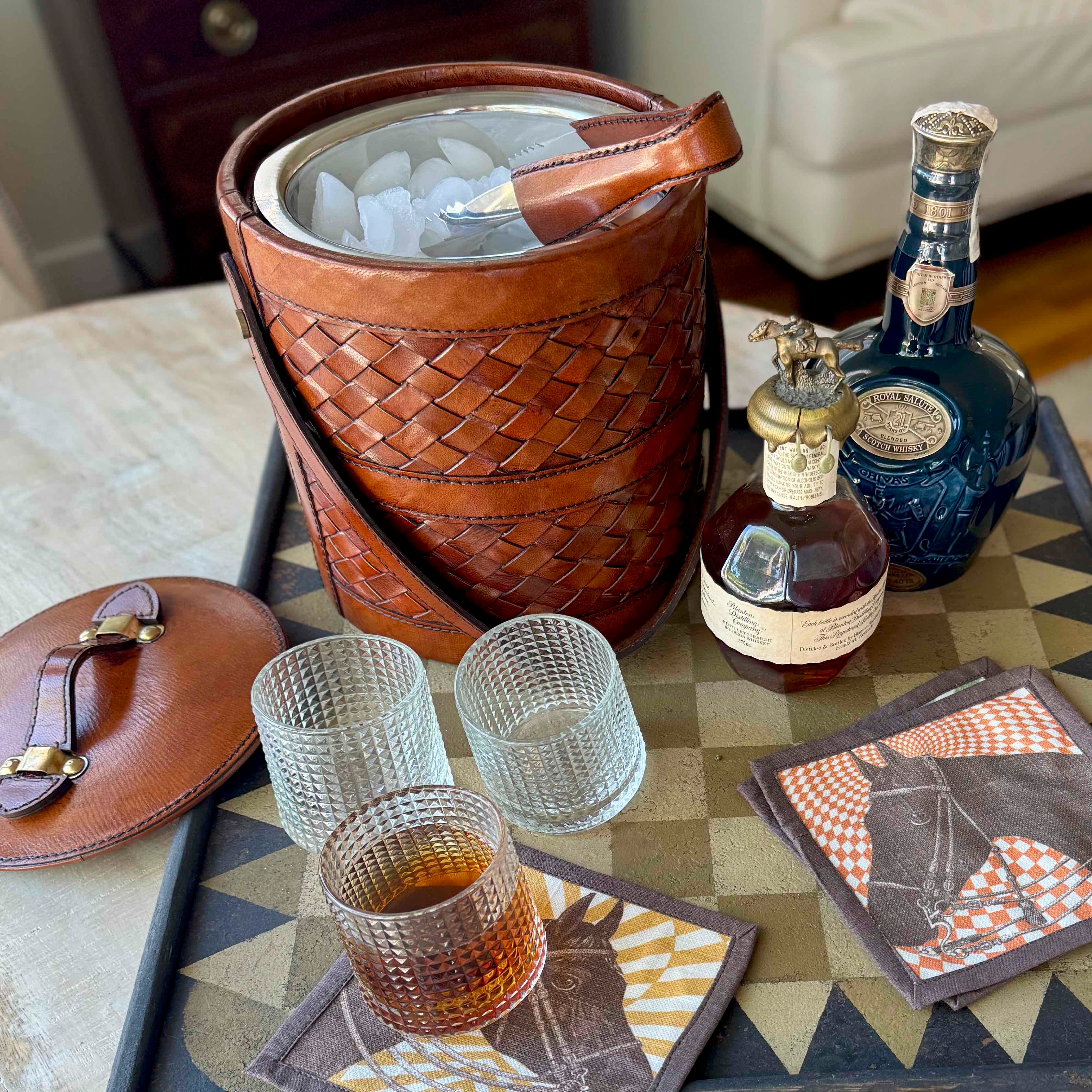 Beverage setup with a woven ice bucket, glasses, bottles, and coasters on a patterned surface.