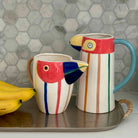 Two colorful ceramic parrot-shaped mugs on a tray with bananas against a hexagonal tile backsplash.