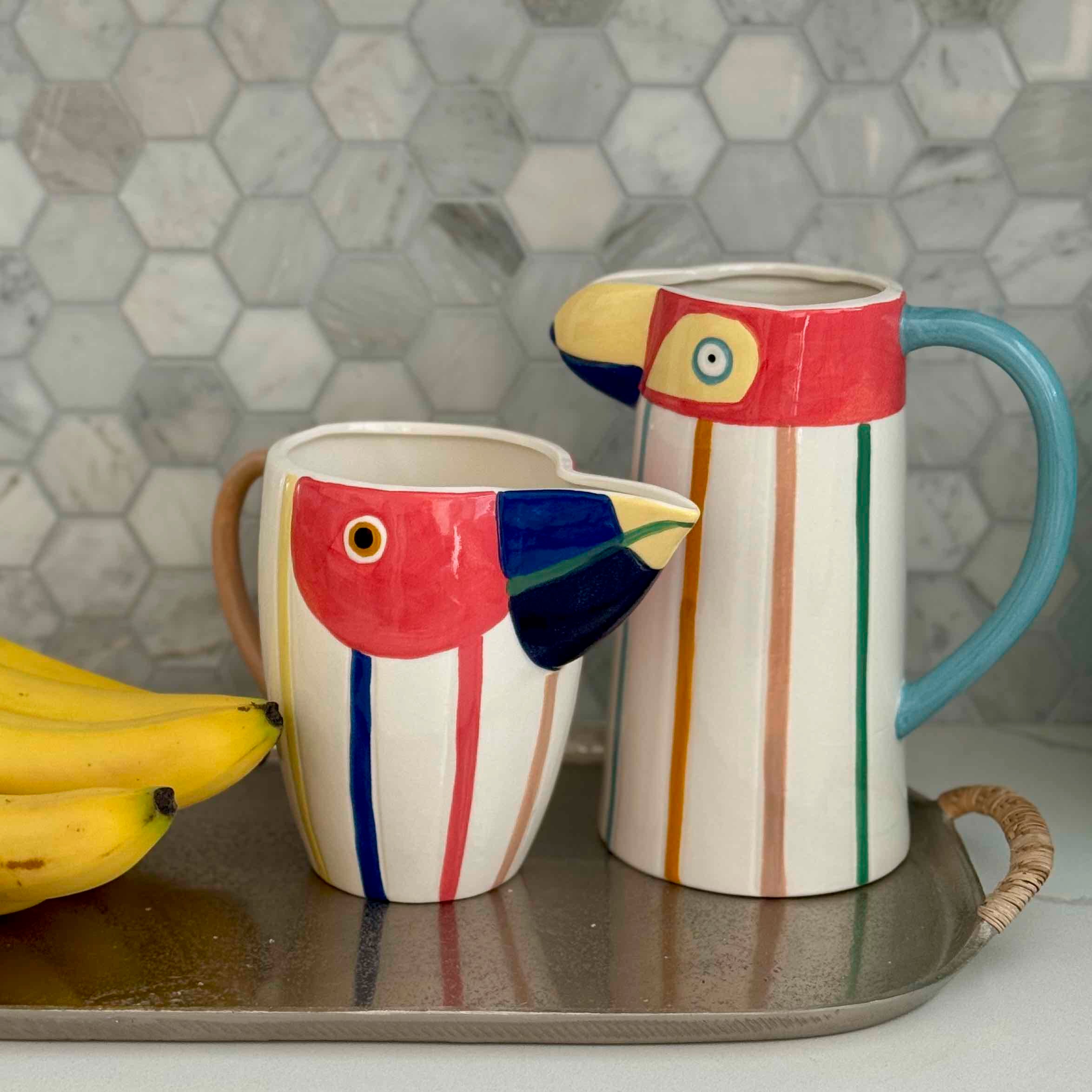 Two colorful ceramic parrot-shaped mugs on a tray with bananas against a hexagonal tile backsplash.
