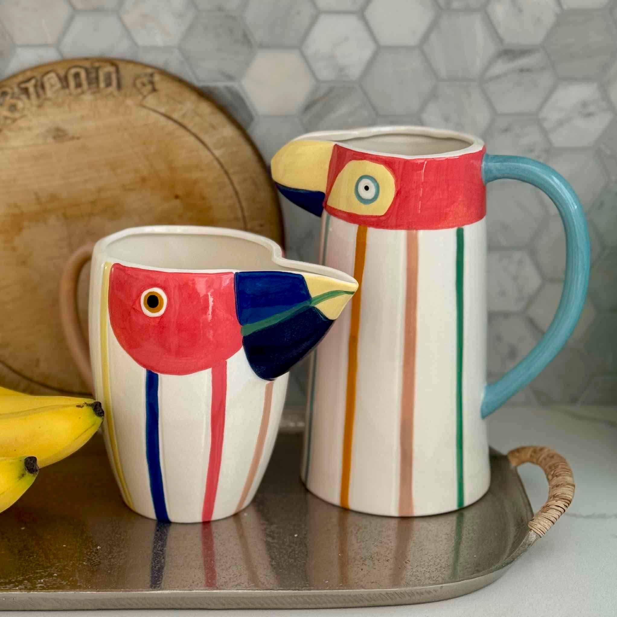 Two colorful ceramic mugs shaped like birds on a tray with a tiled wall background.