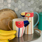 Colorful bird-shaped mugs on a tray with bananas and a hexagonal tile backsplash.