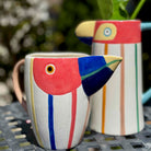 Two colorful ceramic bird-shaped mugs on a metal surface with a blurred natural background.