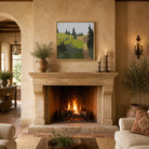 A framed landscape art print hangs on a beige plaster wall above a stone fireplace in a warm, rustic living room. The print depicts rolling green hills and dark pine trees under a soft gray sky.